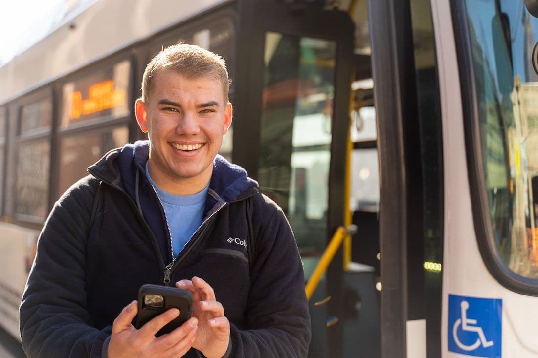 Young man smiling while getting off bus, his mobile device is in his hand.
